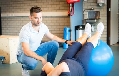 Fysiotherapeut helpt iemand met een ligoefening, waarbij de benen op een gymbal liggen
